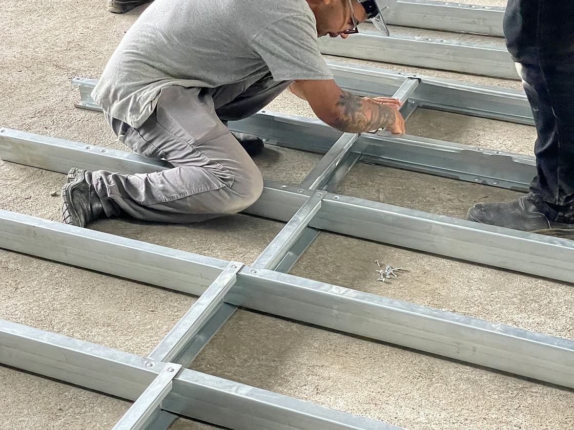 Montador ajustando o painel de piso em perfis de aço galvanizado sobre a laje, com acompanhamento da equipe técnica